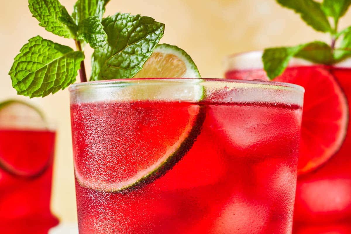 close up of hibiscus iced tea in a glass garnished of mint and lime with two more glasses of hibiscus tea in the background.