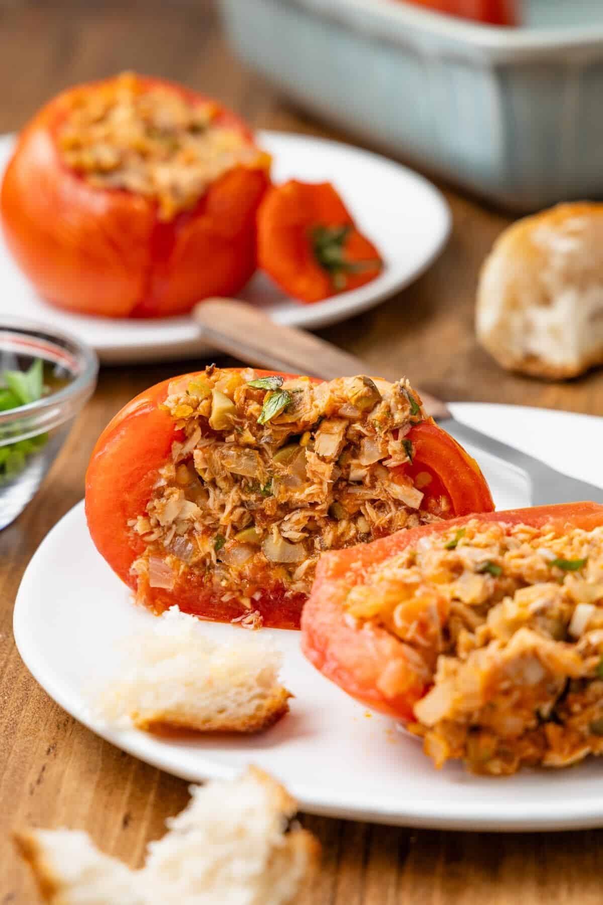 close up of a tomato stuffed with tuna cut in half on a white plate with a fork and a small piece of bread.