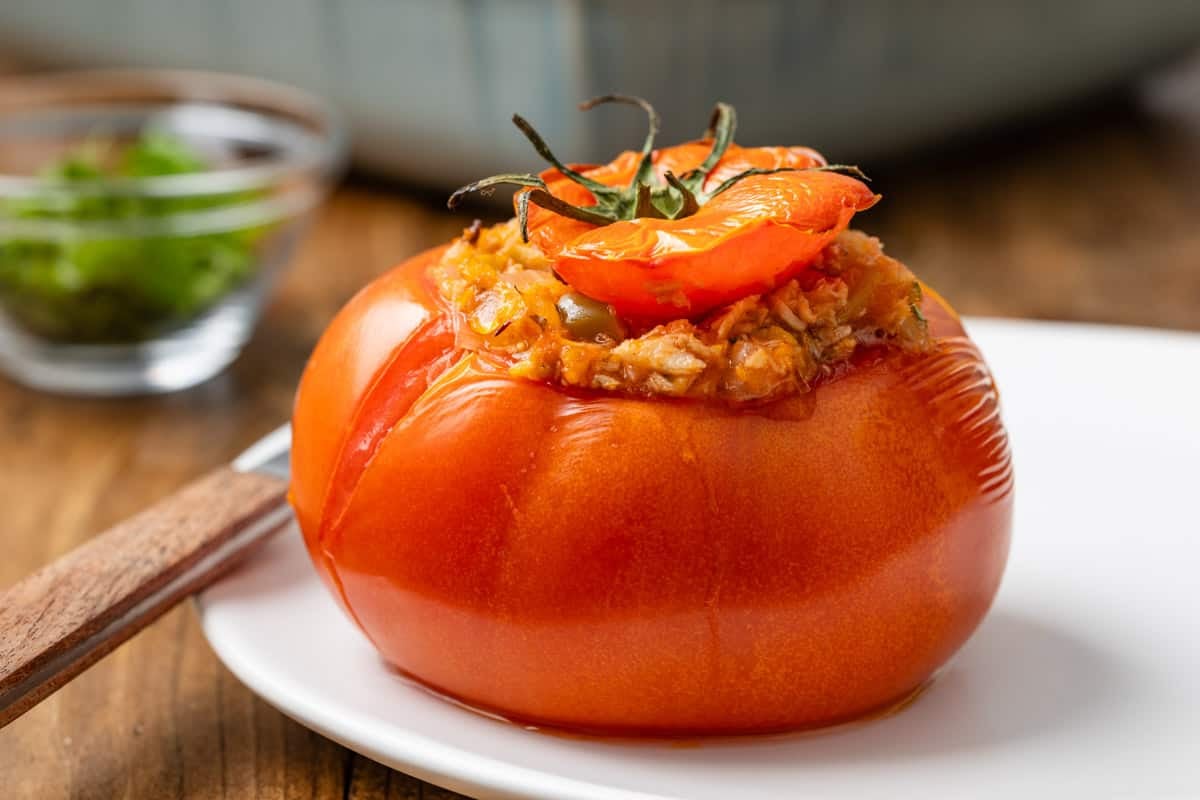 a close up of a baked stuffed tomato with tuna on a white plate with a knife.