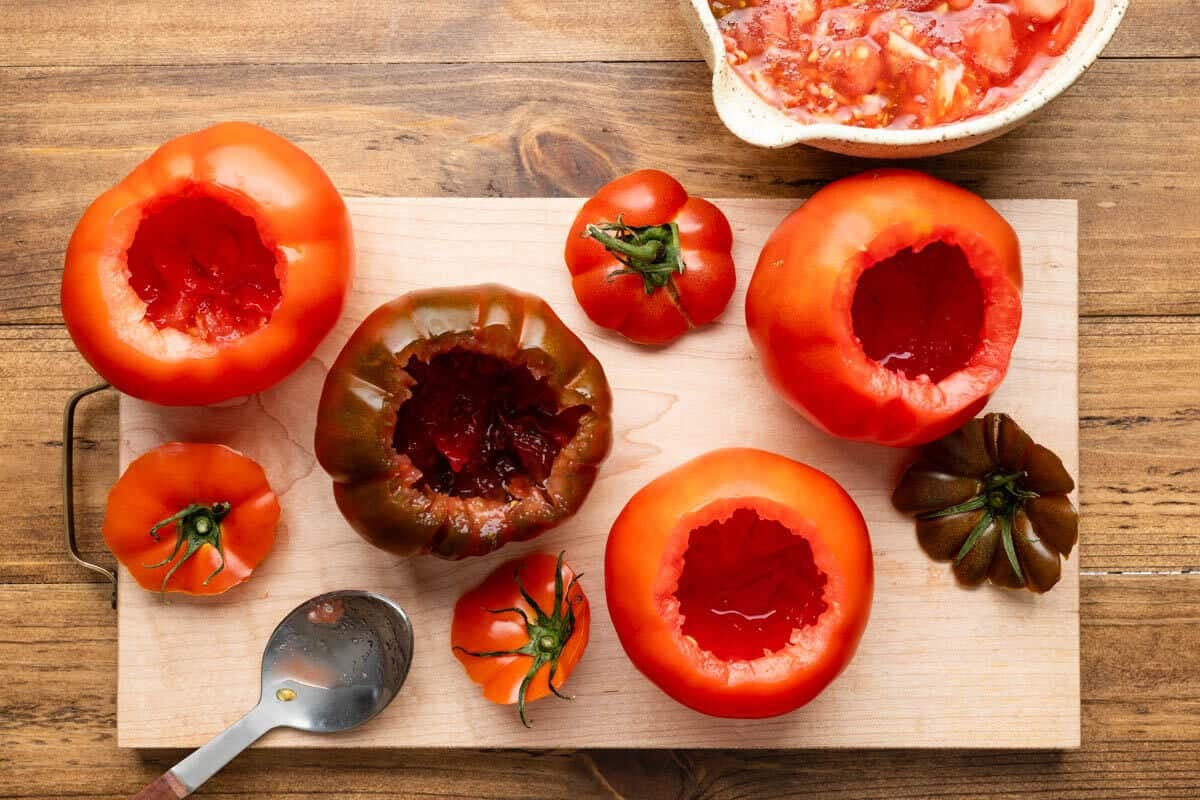 four hollowed out tomatoes on a cutting board next to their tops and a bowl full of tomato flesh.