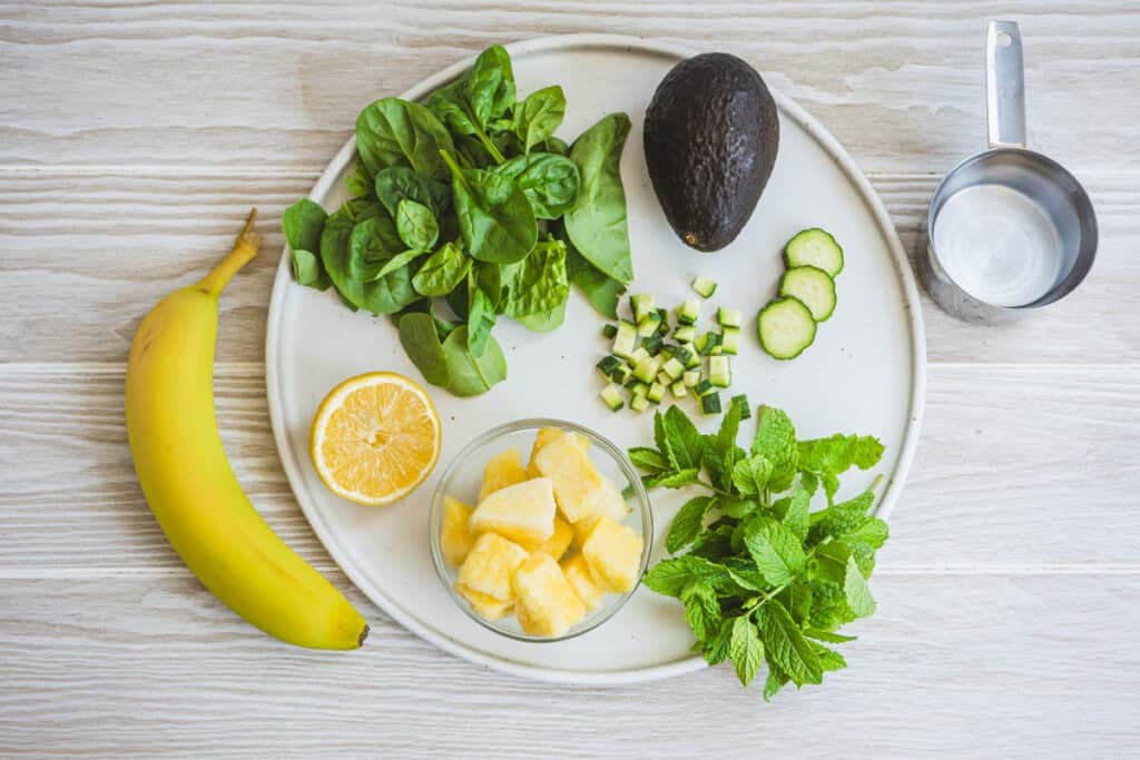 ingredients for a vegan green smoothie with mint including spinach, water, avocado, cucumber, banana, pineapple, lemon, and fresh mint.