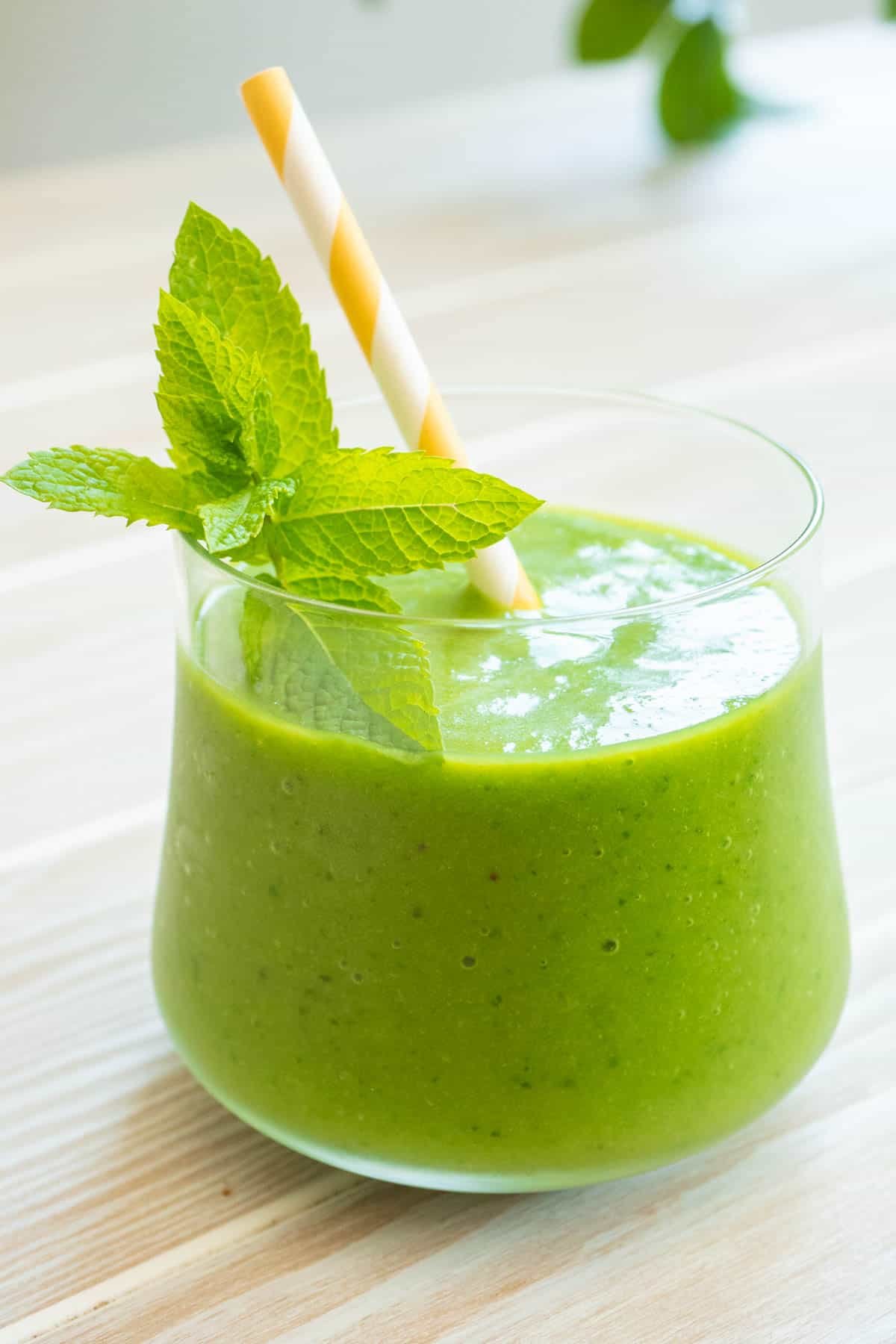 close up of a vegan green smoothie in a glass with mint and a yellow and white straw.