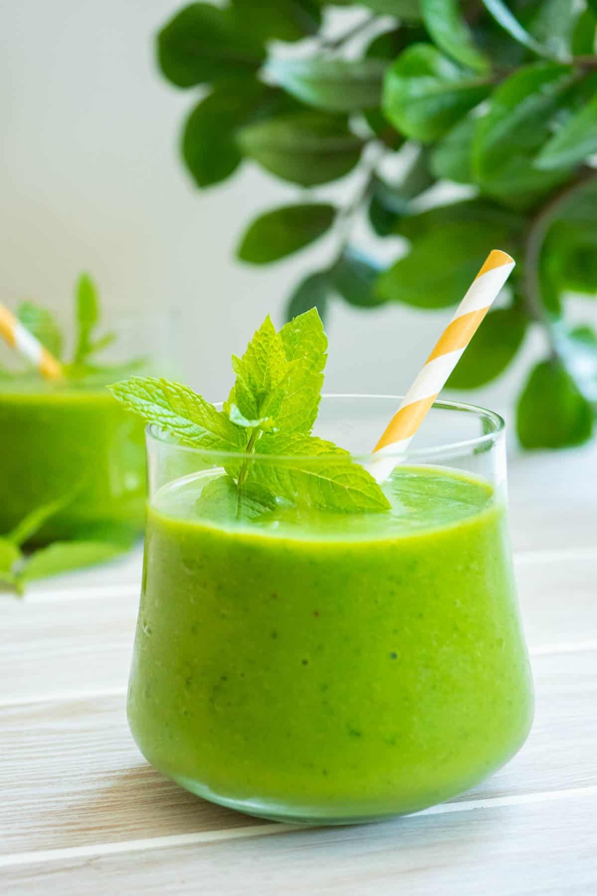a vegan green smoothie in a glass with mint and a yellow and white straw.