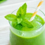 a vegan green smoothie in a glass with fresh mint and a yellow and white straw.