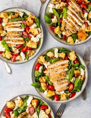 4 chicken caprese salads topped with basil, cherry tomatoes, mozzarella cheese and croutons in bowls with forks next to them.