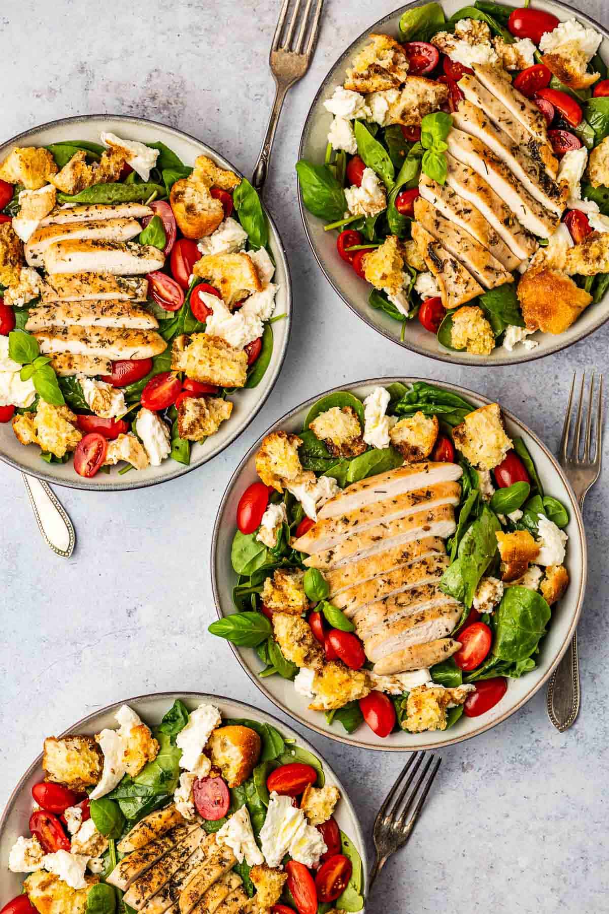 4 chicken caprese salads topped with basil, cherry tomatoes, mozzarella cheese and croutons in bowls with forks next to them.