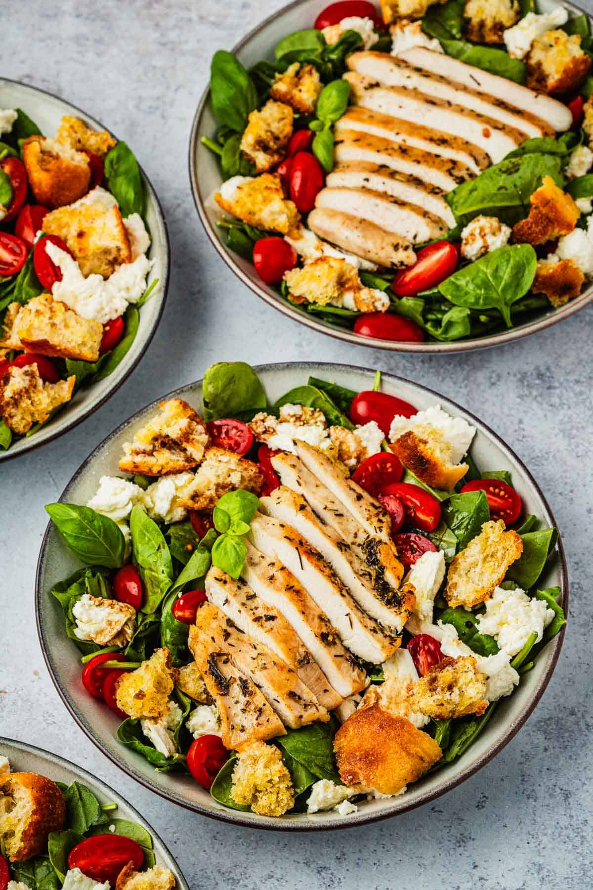 a close up of 4 chicken caprese salads topped with basil, cherry tomatoes, mozzarella cheese and croutons in bowls.