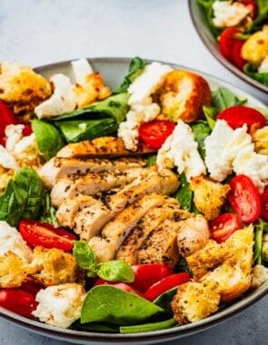 close up of a chicken caprese salad topped with basil, cherry tomatoes, mozzarella cheese and croutons in a bowl.