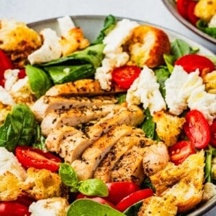 close up of a chicken caprese salad topped with basil, cherry tomatoes, mozzarella cheese and croutons in a bowl.