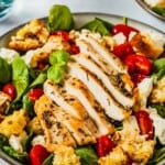 a chicken caprese salad topped with basil, cherry tomatoes, mozzarella cheese and croutons in a bowl.