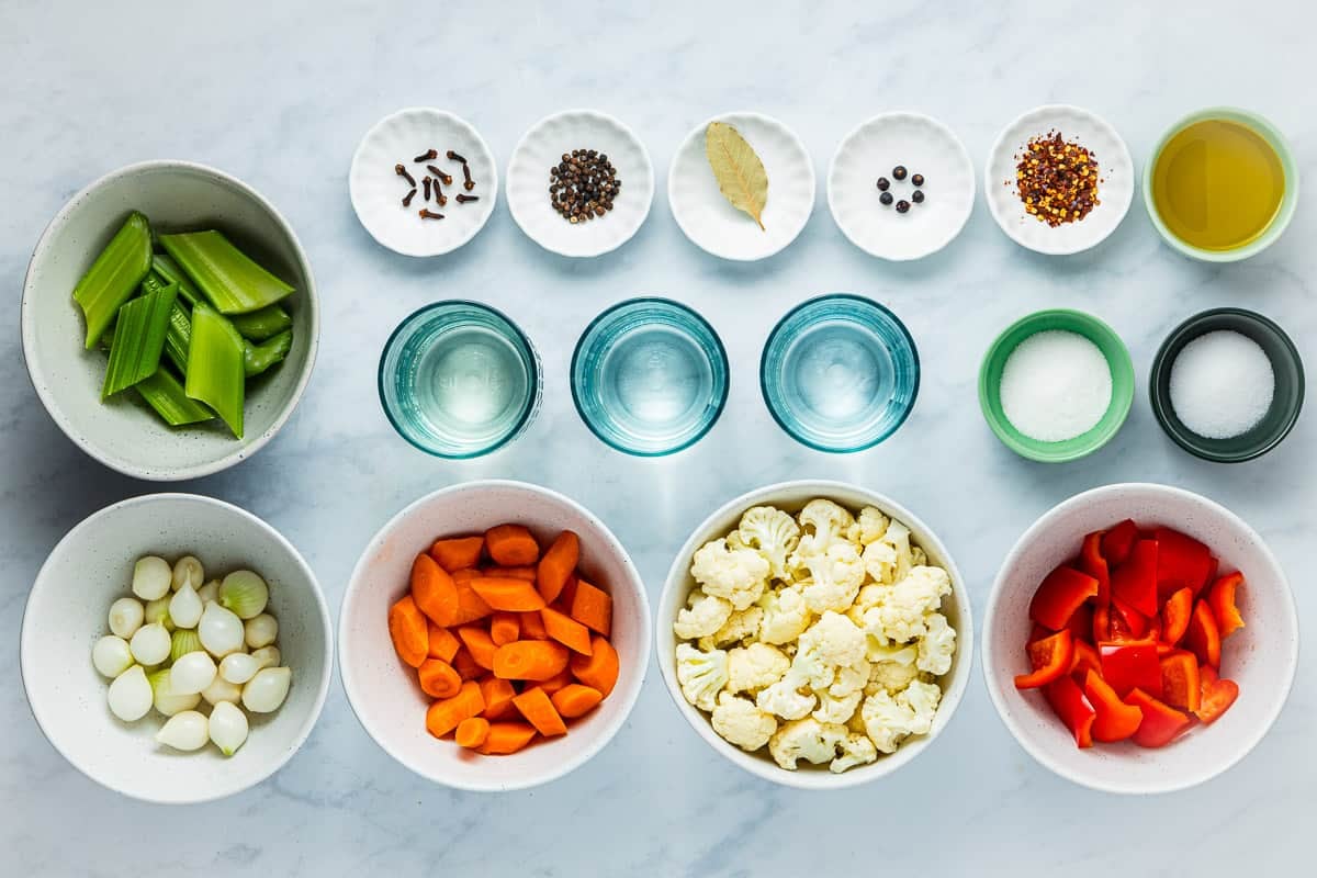 ingredients for giardiniera italian pickled vegetables including olive oil, whole black peppercorns, red pepper flakes, whole cloves, whole juniper berries, a bay leaf, white vinegar, white wine vinegar, water, salt, sugar, cauliflower, carrot, celery, pearl onion and red bell pepper.