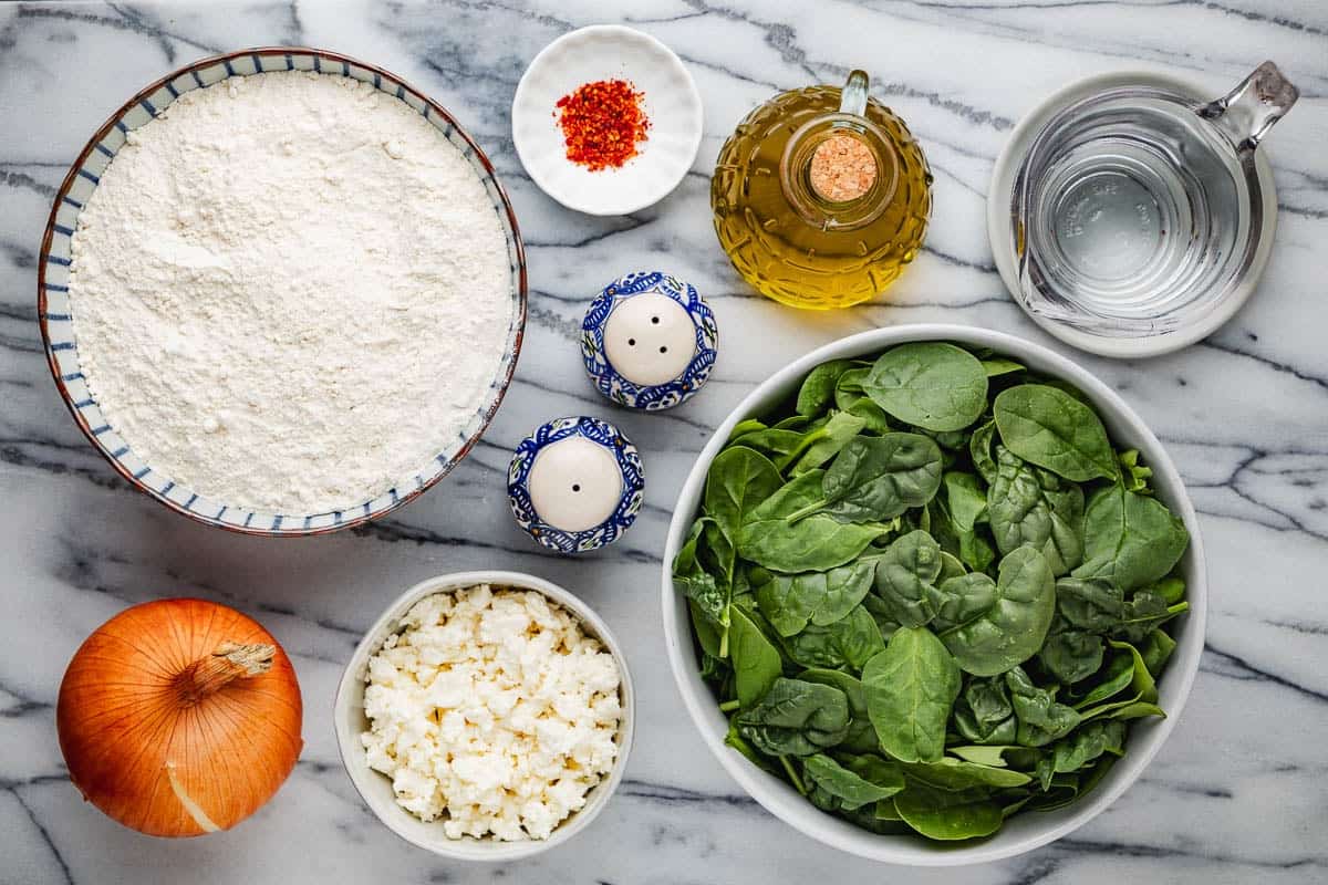 ingredients for gozleme turkish flatbread including all purpose flour, salt, black pepper, water, olive oil, baby spinach, yellow onion, aleppo pepper, and crumbled feta.