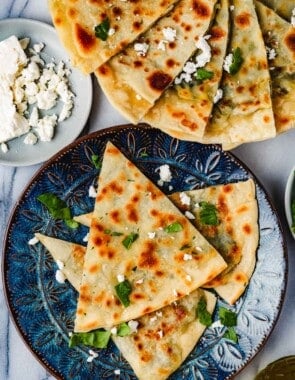 overhead photo of 3 gozleme turkish flatbread triangles on a blue plate and a serving platter with several gozleme triangles next to a plate of crumbled feta.