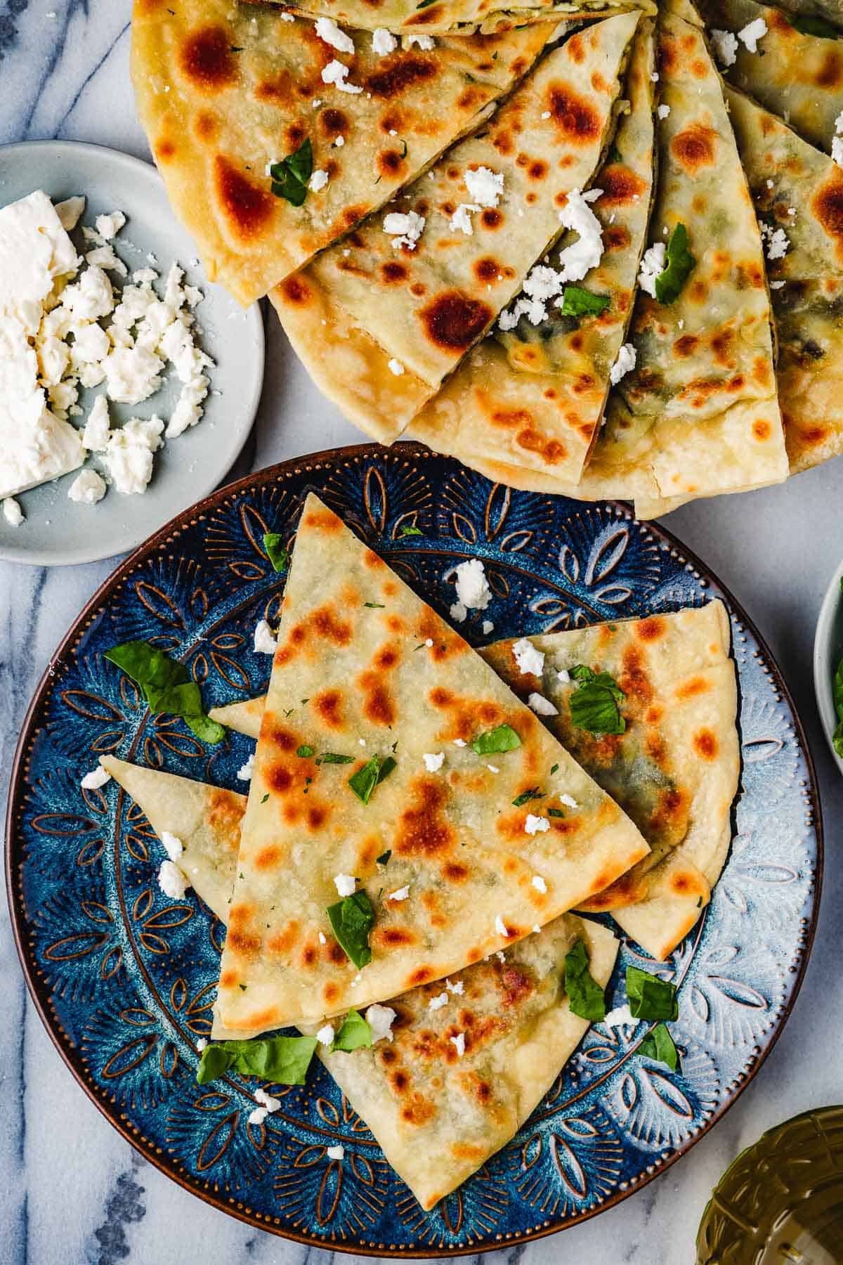 overhead photo of 3 gozleme turkish flatbread triangles on a blue plate and a serving platter with several gozleme triangles next to a plate of crumbled feta.