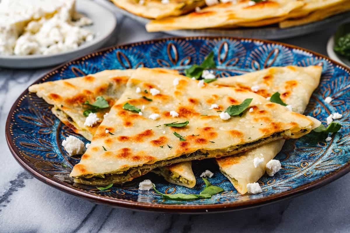 close up of three gozleme turkish flatbread triangles sprinkled with crumbled feta and chopped baby spinach on a blue plate.