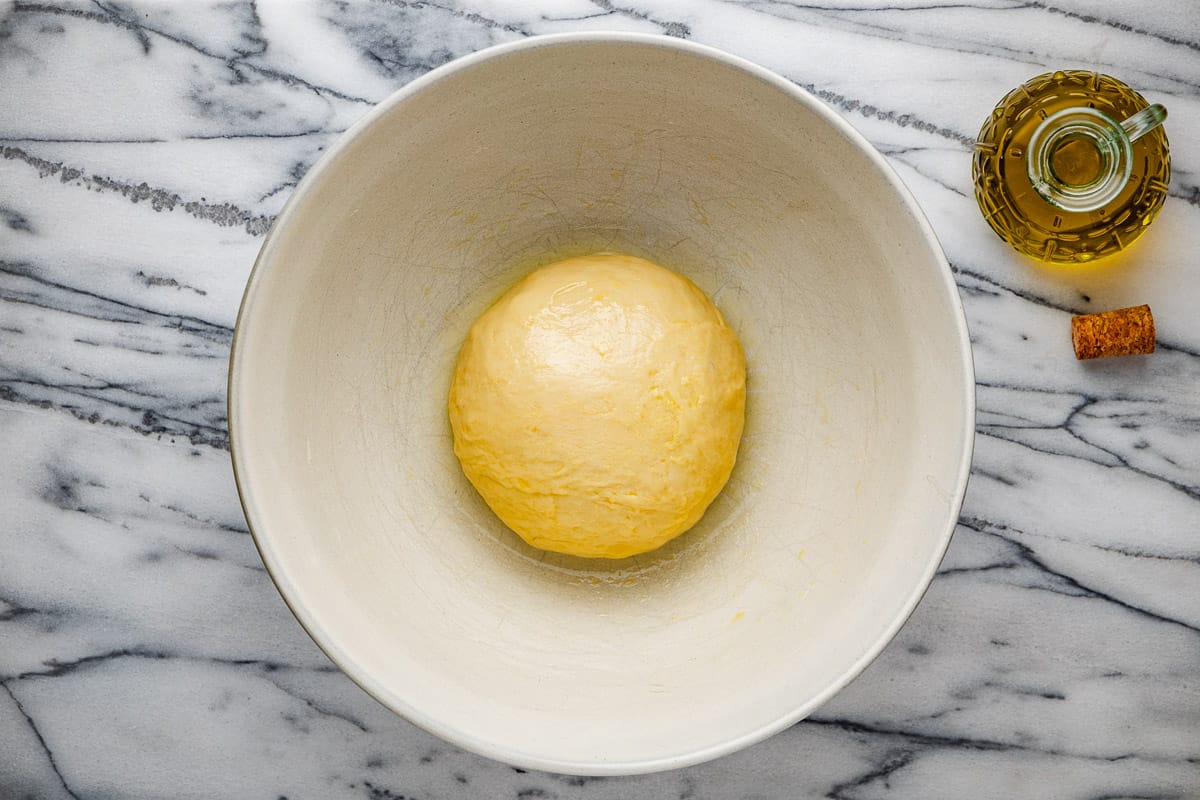 a ball of dough resting in a large white bowl next to a small jug of olive oil.