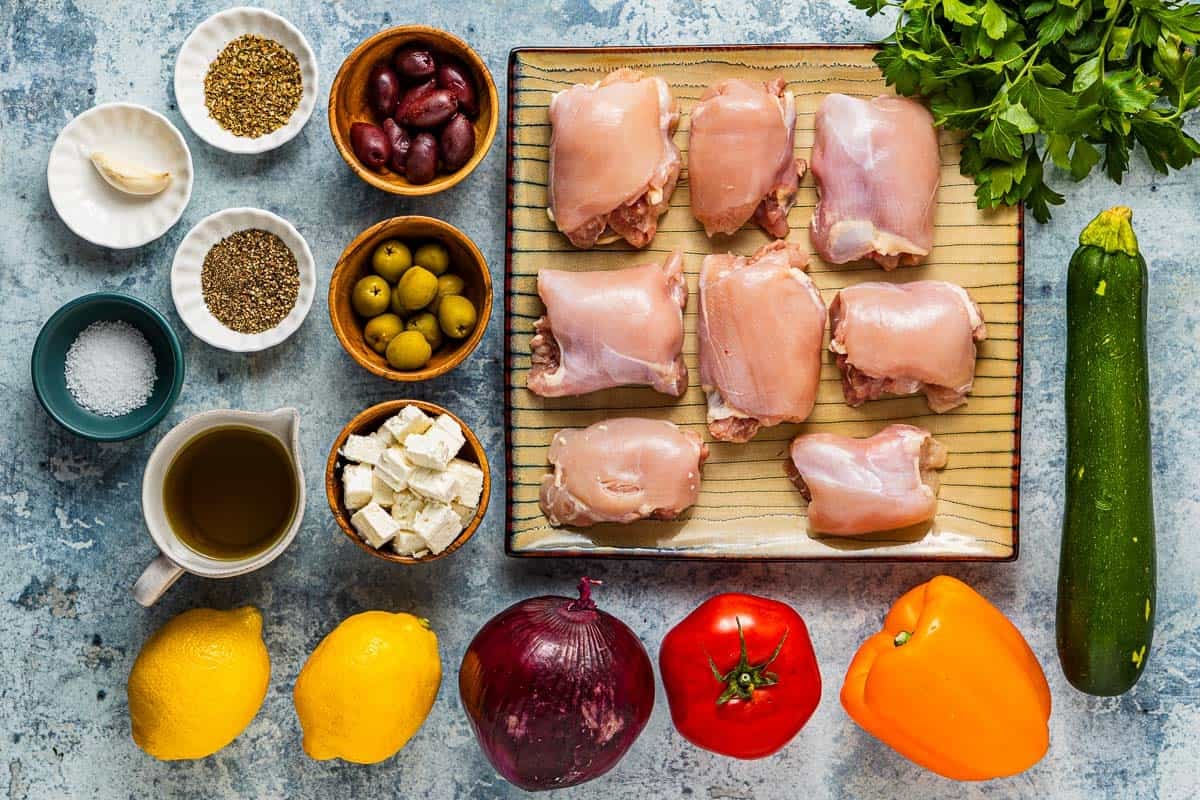ingredients for Greek sheet pan chicken including chicken thighs, lemons, tomato, red onion, zucchini, orange bell pepper, parsley, kalamata olives, green olives, feta cheese, garlic, oregano, salt, pepper and olive oil.