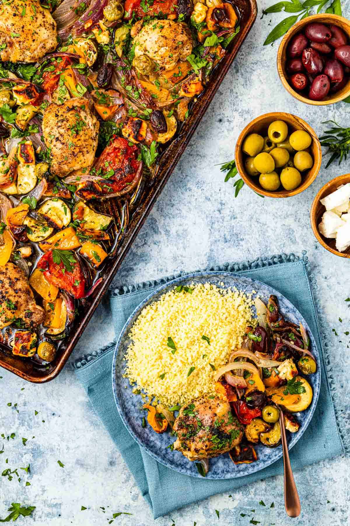 baked Greek sheet pan chicken and vegetables on a plate with a side of couscous, next to a full sheet pan of Greek chicken and vegetables and bowls of green olives, kalamata olives and feta cheese.