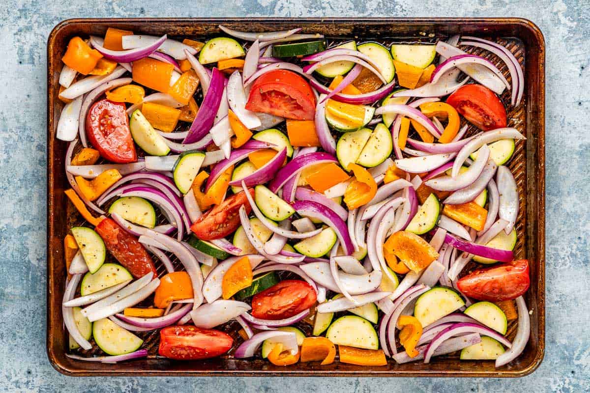 slices of uncooked tomatoes, red onion, zucchini, and orange bell pepper on a sheet pan.