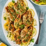 pieces of lemon garlic chicken garnished with lemon wheels on a white serving platter next to a fork and a side salad in a bowl.