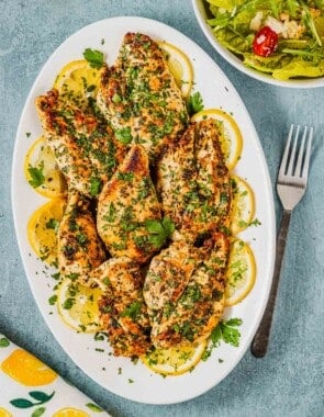 pieces of lemon garlic chicken garnished with lemon wheels on a white serving platter next to a fork and a side salad in a bowl.