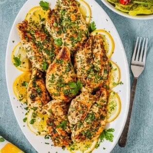 pieces of lemon garlic chicken garnished with lemon wheels on a white serving platter next to a fork and a side salad in a bowl.