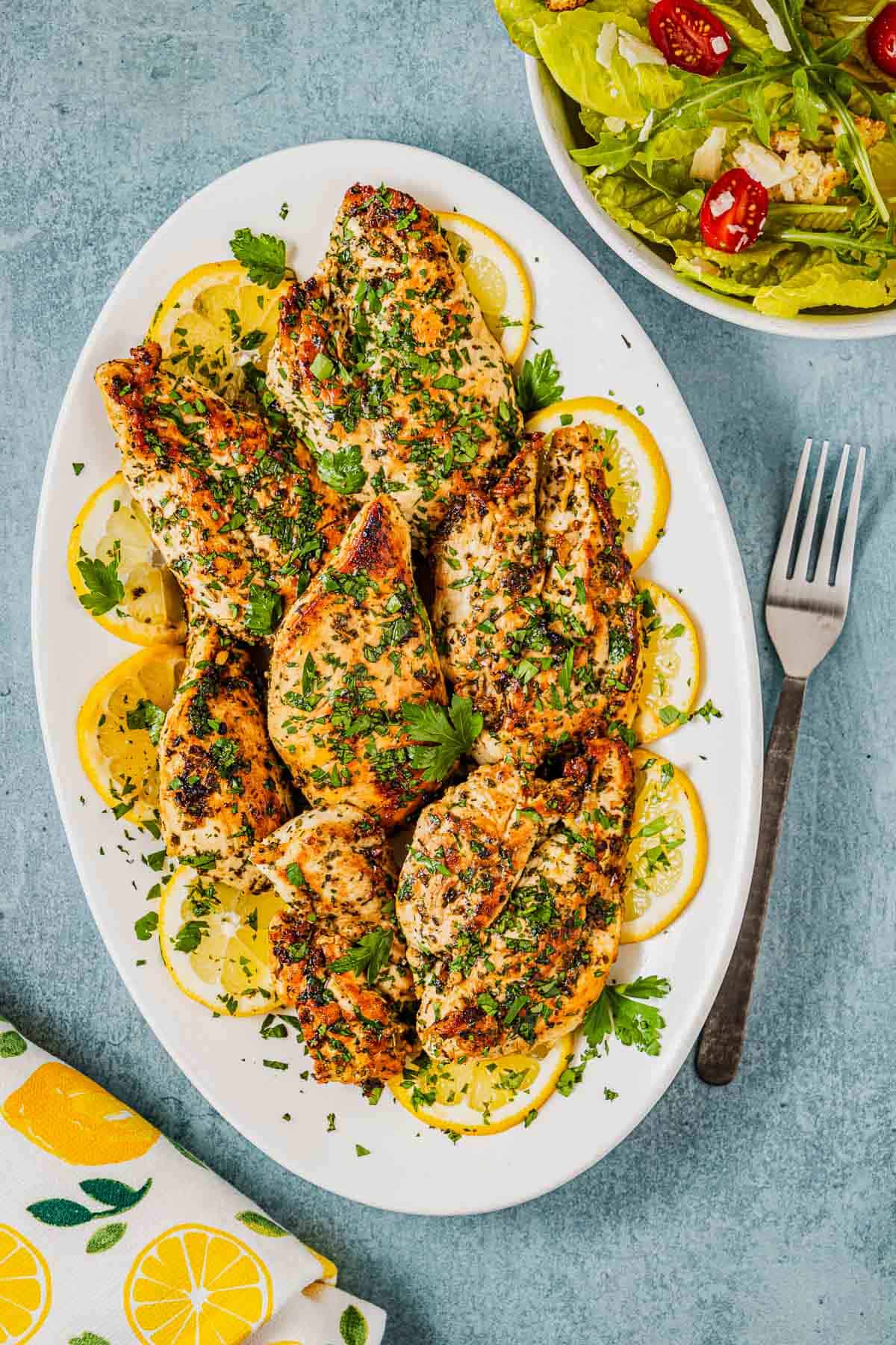 pieces of lemon garlic chicken garnished with lemon wheels on a white serving platter next to a fork and a side salad in a bowl.