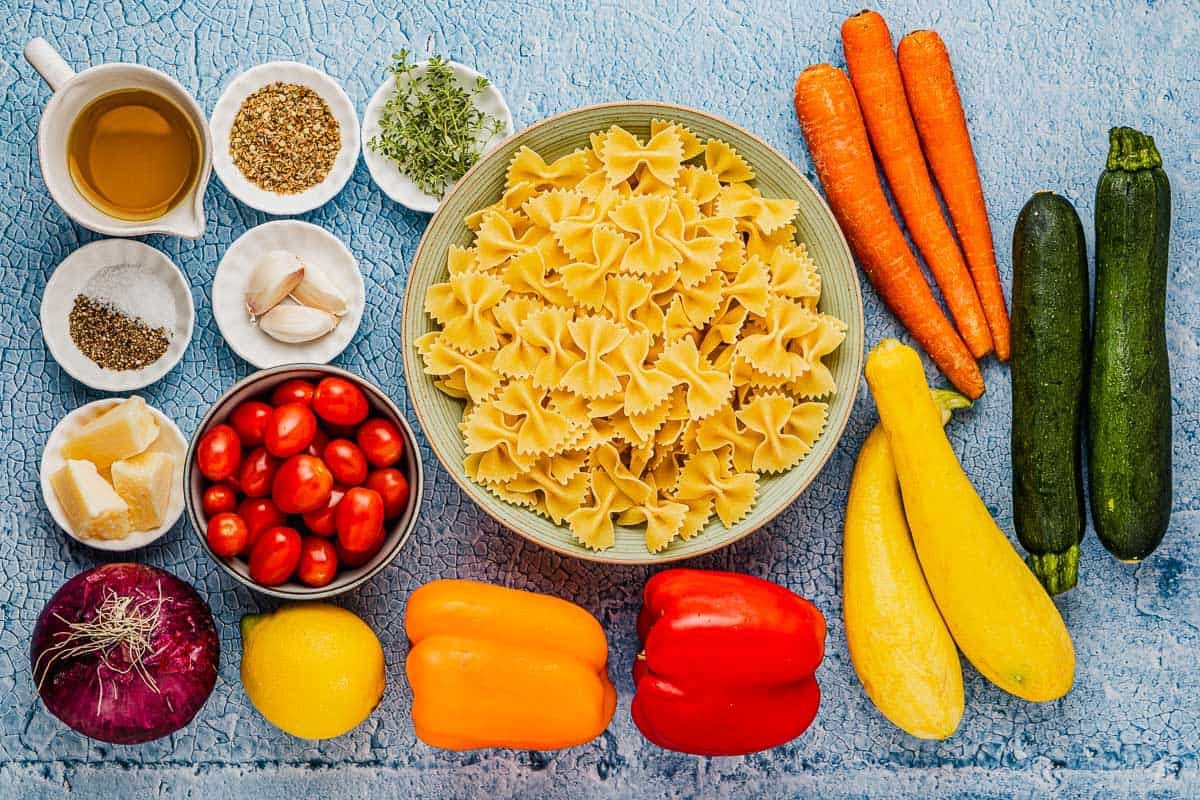 ingredients for pasta primavera including bow tie pasta, carrots, zucchini, yellow squash, orange bell pepper, red bell pepper, lemon, red onion, cherry tomatoes, parmesan cheese, garlic, salt, pepper, olive oil, oregano, and thyme.