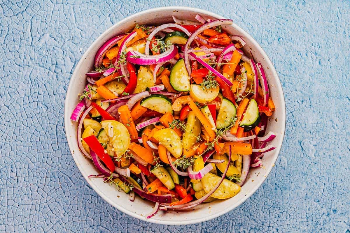 pieces of zucchini, yellow squash, carrots, red bell pepper, yellow bell pepper, red onion plus salt, pepper, garlic, oregano, thyme and a drizzle of olive oil in a white mixing bowl.