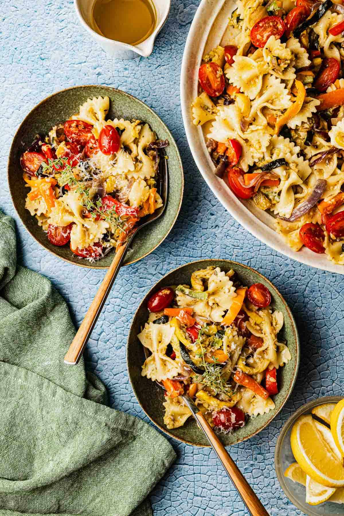 two green bowls of pasta primavera with forks next to a serving bowl with pasta primavera, a cup of olive oil and a bowl of lemon wedges.