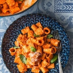 a blue plate with creamy roasted red pepper pasta topped with parmesan cheese and basil with a fork next to a pot of roasted red pepper pasta.