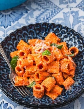 a blue plate with creamy roasted red pepper pasta topped with parmesan cheese and basil with a fork.