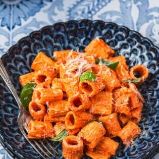 a blue plate with creamy roasted red pepper pasta topped with parmesan cheese and basil with a fork.