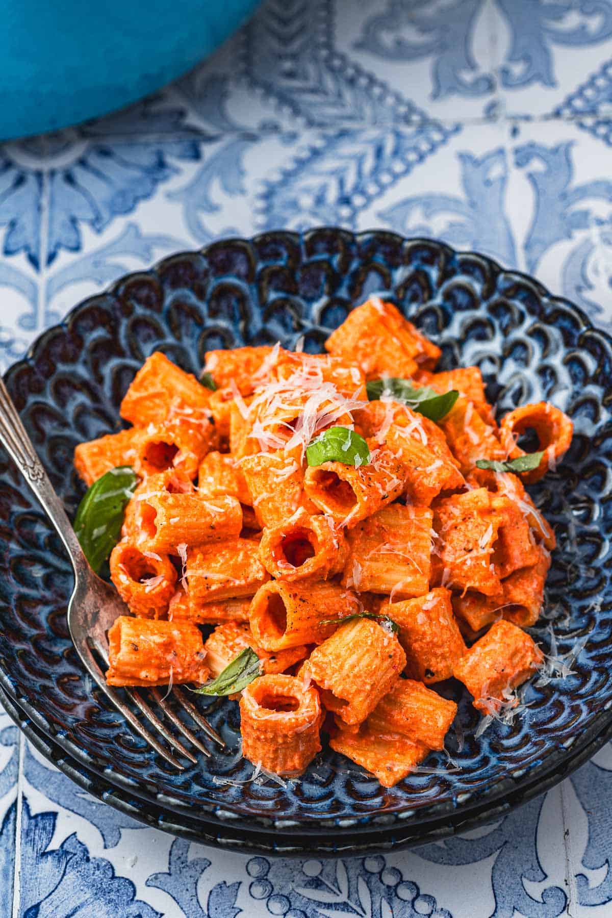 a blue plate with creamy roasted red pepper pasta topped with parmesan cheese and basil with a fork.