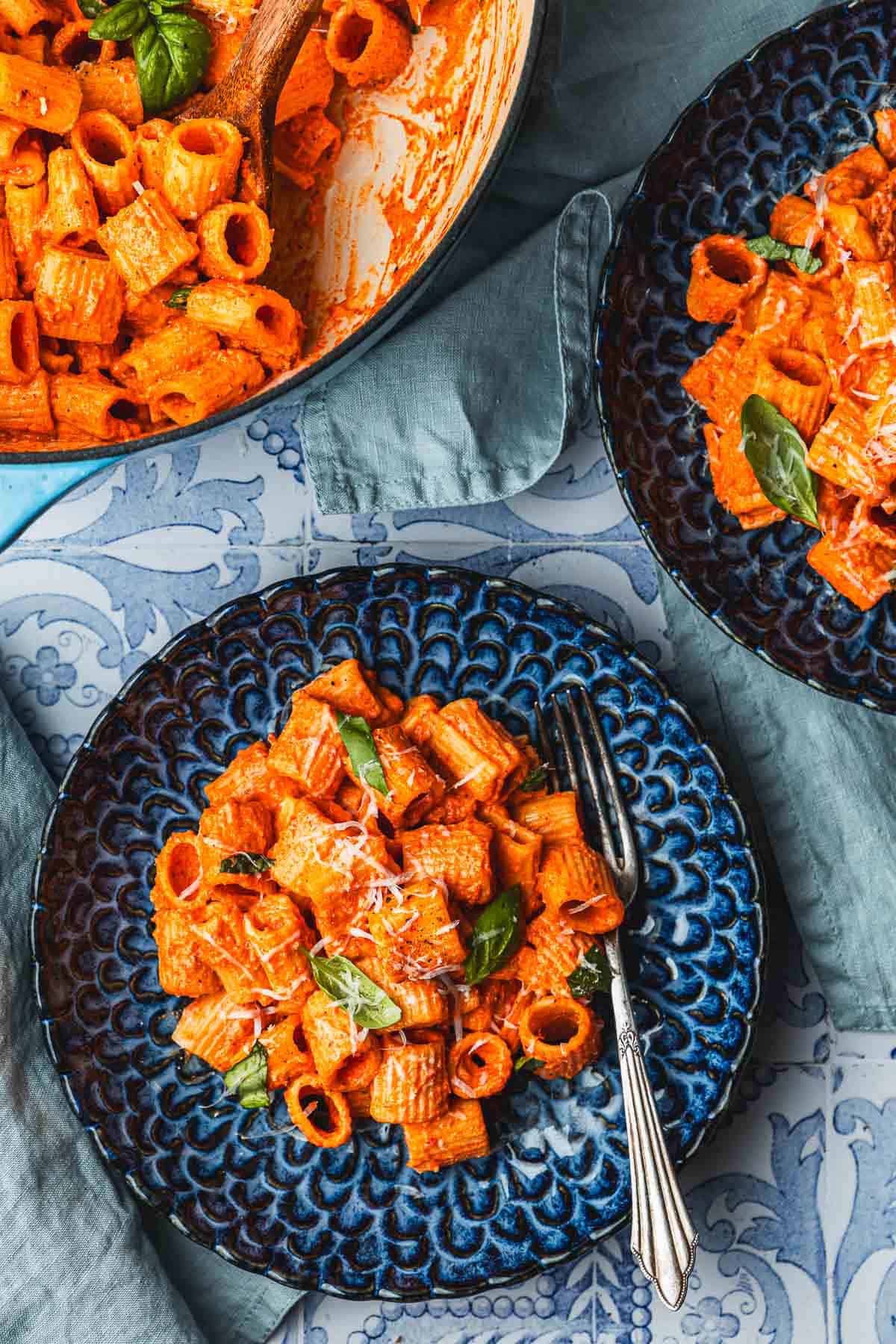 two plates with creamy roasted red pepper pasta topped with parmesan cheese and basil next to a pot of roasted red pepper pasta.