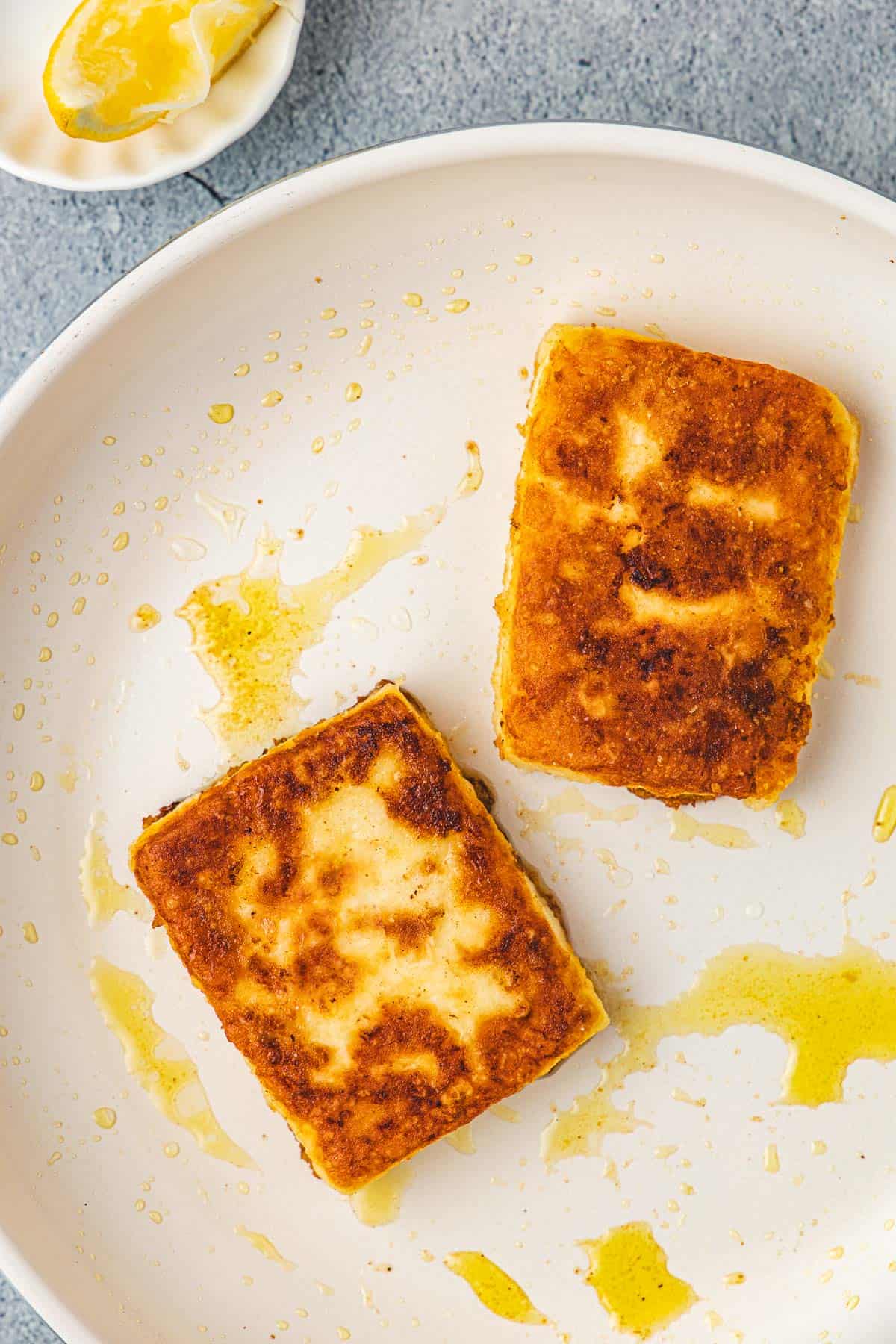 overhead photo of two slices of saganaki fried greek cheese in a skillet next to a bowl with a lemon wedge.