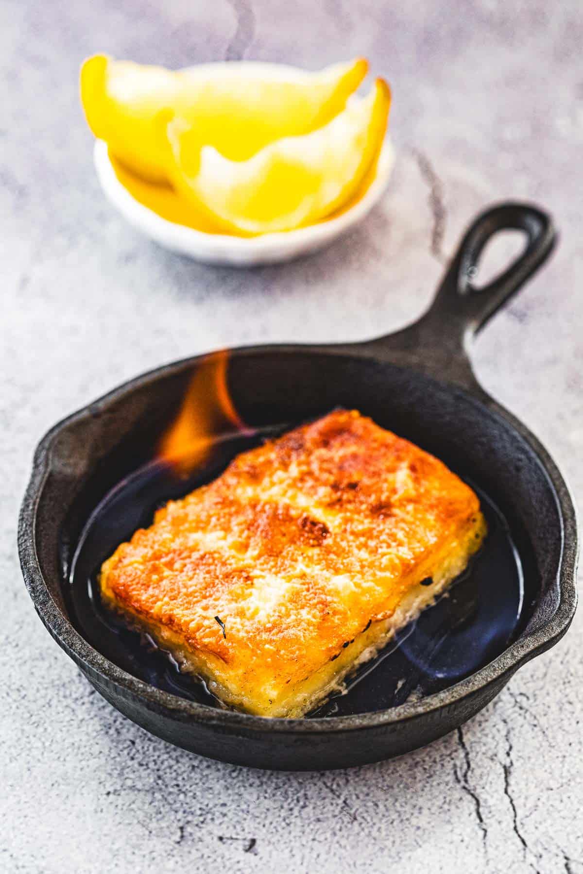 one flaming saganaki fried greek cheese in a mini cast iron skillet next to a bowl with 2 lemon wedges.