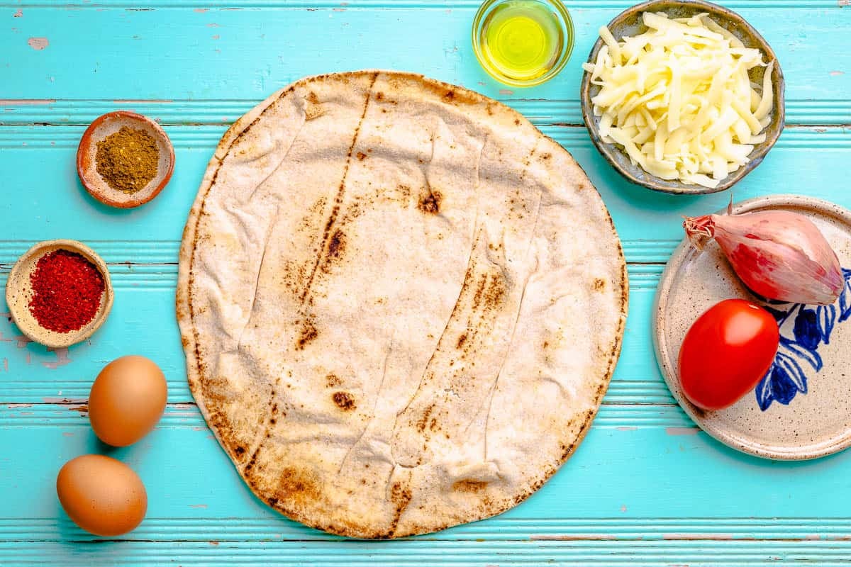 ingredients for breakfast pizza including a large whole wheat pita, olive oil, grated mozzarella, za'atar, red pepper flakes, 1 shallot, 1 roma tomato, and 2 hard boiled eggs.