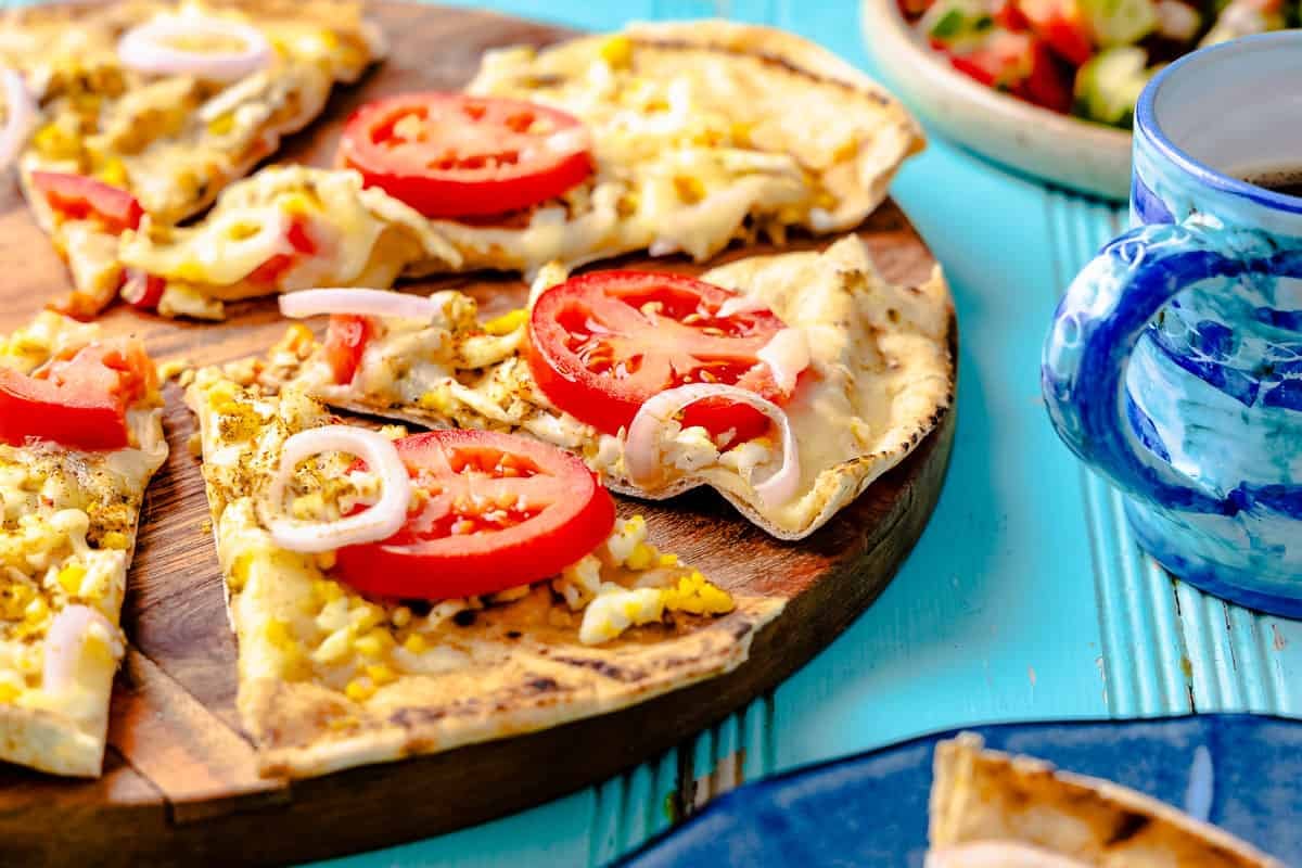 a pita breakfast pizza on a wooden serving platter sliced into individual pieces next to a cup of coffee.