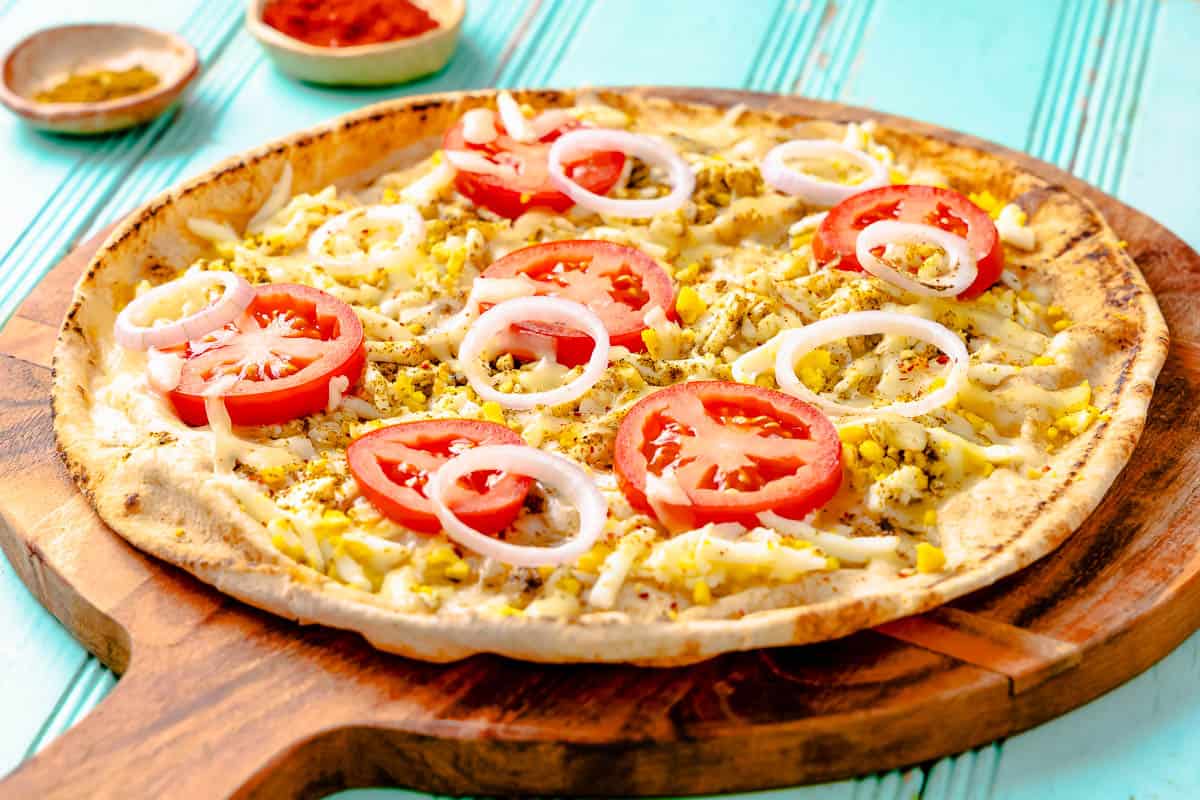 close up of a baked pita breakfast pizza on a wooden serving platter next to bowls of za'atar and red pepper flakes.