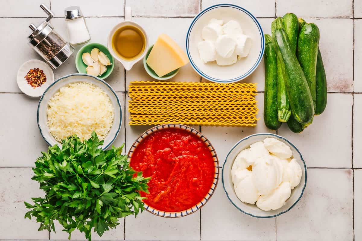 ingredients for lasagna roll ups including zucchini, lasagna noodles, ricotta cheese, goat cheese, mozzarella cheese, parmesan cheese, parsley, garlic cloves, salt, pepper, red pepper flakes, pasta sauce and olive oil.