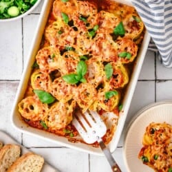 baked lasagna roll ups in a baking dish with a spatula topped with basil leaves next to a bowl of salad, a plate of bread, and another plate with 2 lasagna roll ups.