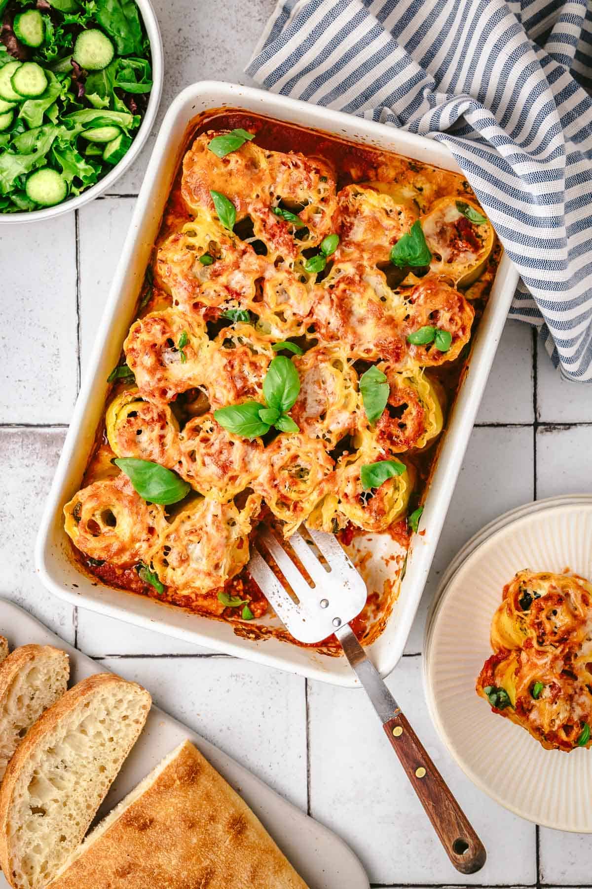 baked lasagna roll ups in a baking dish with a spatula topped with basil leaves next to a bowl of salad, a plate of bread, and another plate with 2 lasagna roll ups.