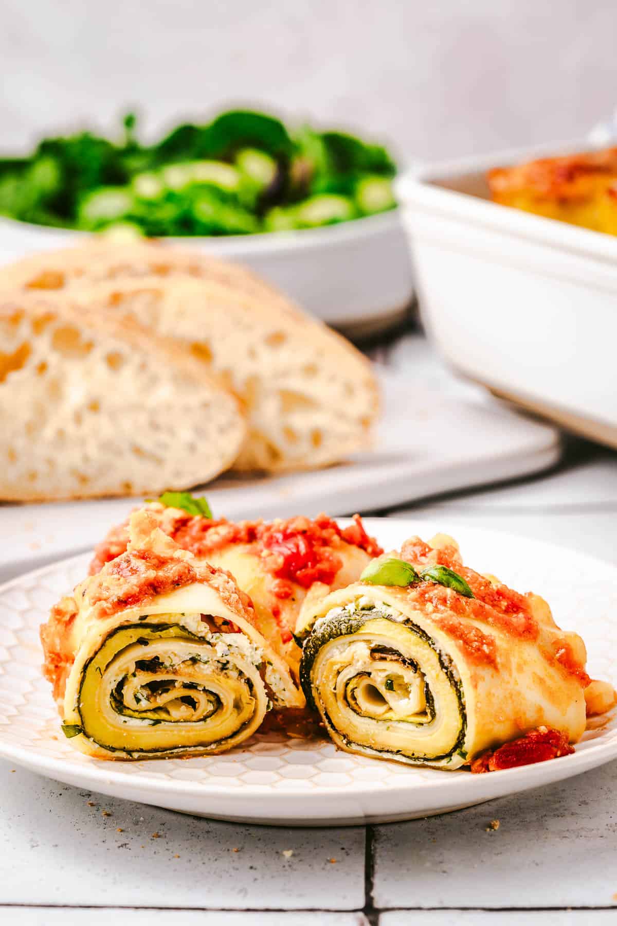 a lasagna roll up cut in half on a plate with a plate of sliced bread and a bowl of salad in the background.