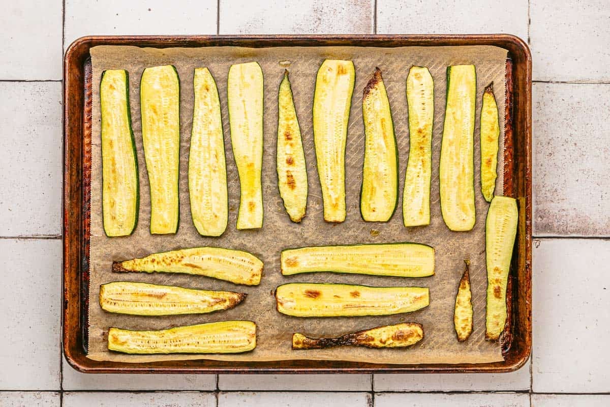 several baked slices of zucchini arranged on a parchment lined baking sheet.