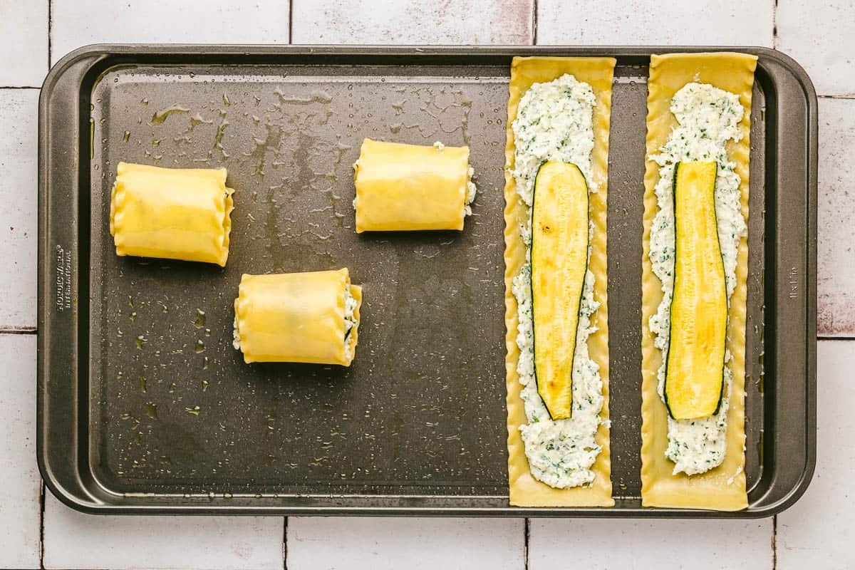 5 lasagna noodles with a cheese filling and a slice of zucchini on a sheet pan, three rolled up and two unrolled.