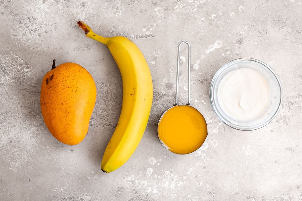 Ingredients for a mango smoothie, including one mango, one banana, orange juice, and yogurt.