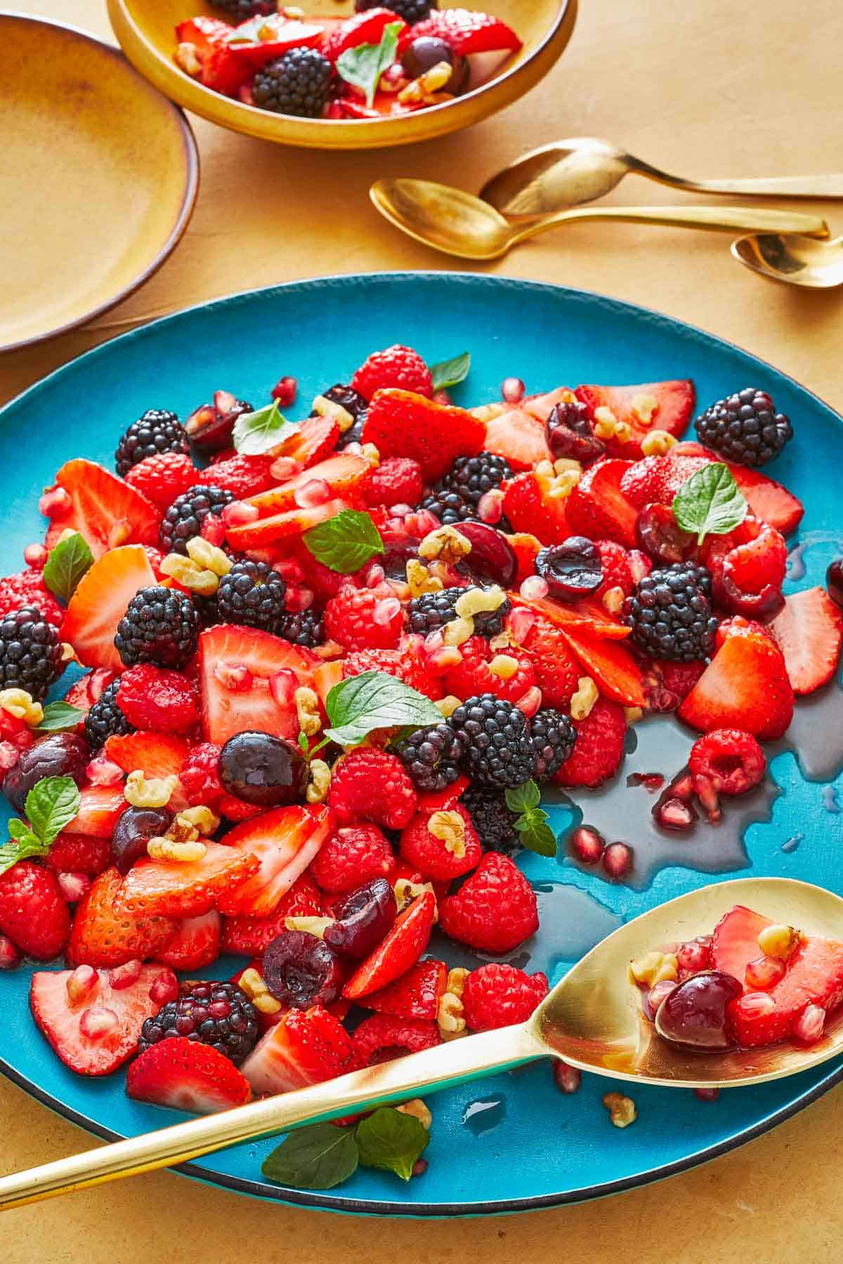 fruit salad topped with mint and walnuts on a blue plate with a gold serving spoon next to 3 gold teaspoons, a small bowl of fruit salad and an empty plate.