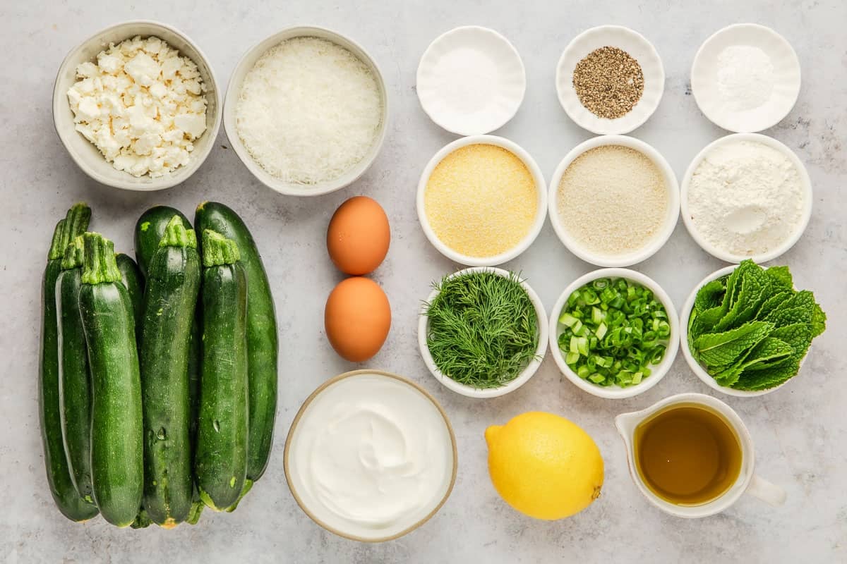 Ingredients for zucchini fritters, including feta cheese, zucchini, mint, dill, salt, pepper, lemon, olive oil, two eggs, green onion, parmesan cheese, breadcrumbs, all purpose flour, and semolina flour.