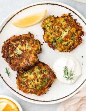 Three zucchini fritters on a plate with a dollop of Greek yogurt and a lemon wedge.